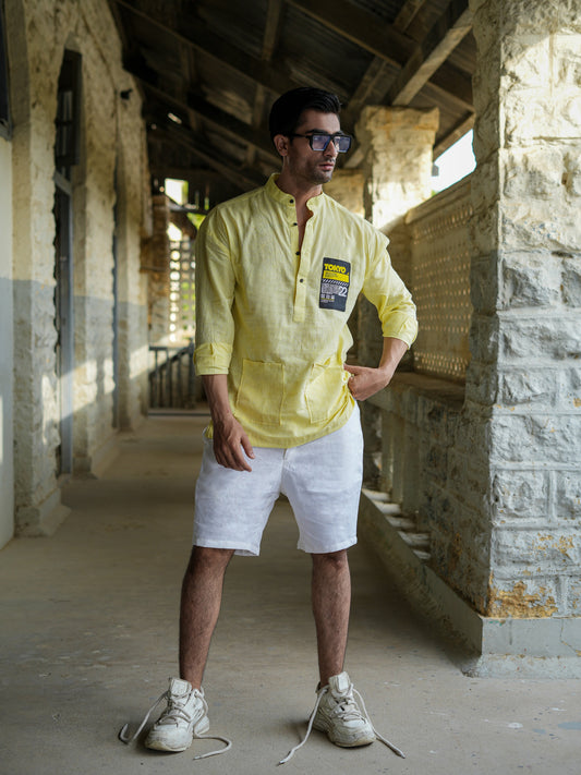 Man wearing a yellow shirt with a logo and white shorts standing in a sunlit hallway.